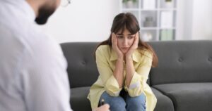 - Addiction Free Recovery Young woman sits on a gray sofa with her hands pressed to her cheeks, looking sad during a therapy session.